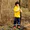 A young boy wearing Muddy Puddles Children's Waterproofs, consisting of a bright yellow raincoat and dark blue waterproof trousers, stands in a woodland.